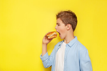 A teenage boy bites a burger wide open his mouth. A boy in a blue shirt, his hair ruffled against a yellow background. Copy space.