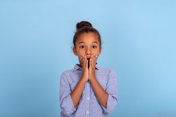 Young girl  coveres her mouth in surprise against a blue background in studio.