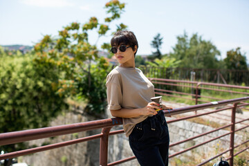 Young woman with short hair and circular sunglasses relaxing with cup of coffee leaning on the fence