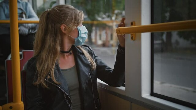 Woman hipster in protective mask is sitting in moving tram and looking out the window. Coronavirus pandemic. Slow motion, dramatic cinematic portrait