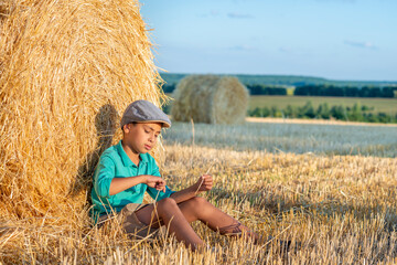Naklejka na ściany i meble One young boy sits at a haystack in a field on a sunny day and thinks about something.