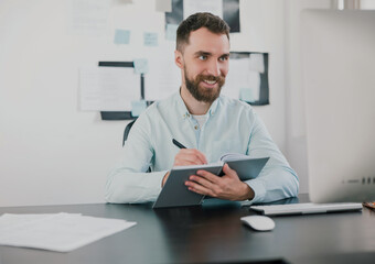 Fototapeta premium young bearded brunette smiling man looking happy while taking notes to his agenda working on business project in his modern office, work routine concept