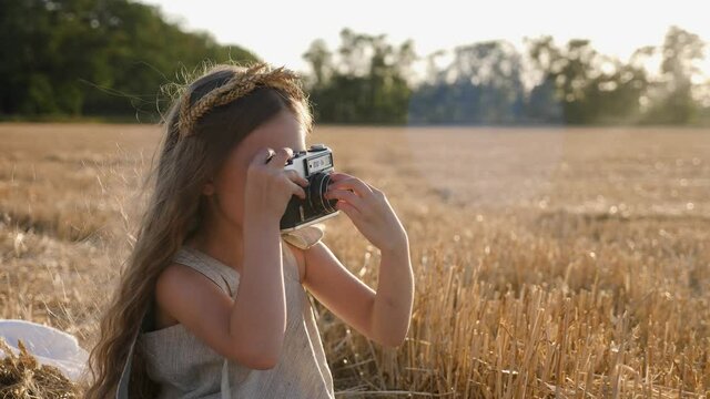 serious girl child in a dress with long hair sits on a mown wheat field and holds a retro camera in her hands