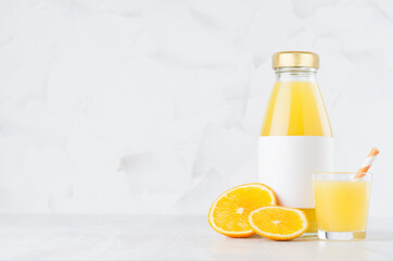 Bright fresh orange juice in glass bottle with blank label template with wine glass decorated straw, fruit slices in soft light white interior on wood table with copy space.