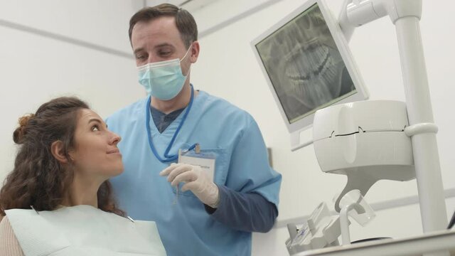 Lockdown of young curly-haired woman lying on dentist chair while professional orthodontist wearing medical uniform examining her teeth. Then they looking at camera