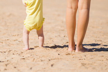Fototapeta na wymiar Young mother and baby legs standing together on sand at beach in sunny summer day. Back view. Closeup.