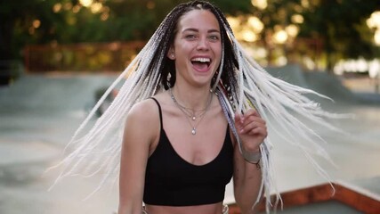 Cheerful girl with dreadlocks dancing in a park. Beautiful woman in black top dancing actively during a sunny day. Smiling to the camera, carefree, turning around. Skatepark blurred on background