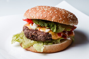 classic burger with a beef patty, cheese, onions, ketchup, tomato slices and salad leaves in a burger bun with sesame on a white plate