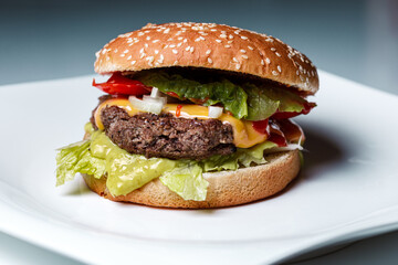 classic burger with a beef patty, melted cheese, onions, ketchup, tomato slices and salad leaves in a burger bun with sesame on a white plate