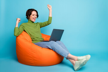 Full size profile side photo of delighted girl sit bean chair work laptop raise fists wear autumn style pullover denim jeans isolated over blue color background