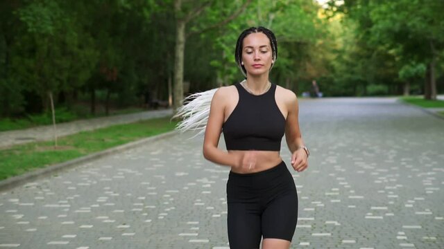 Front view of european woman with black and white dreadlocks running by local, green park in the city. She is exercising for good health. Wearing black top and shorts. Slow motion