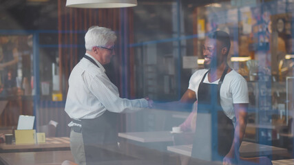 View through window of two diverse owners of coffee shop in aprons shaking hands