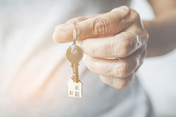old woman's finger hold house key with blurred mini wooden house , close up