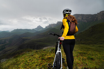 a girl in a helmet and with a backpack with a bicycle stands against the backdrop of epic mountains...