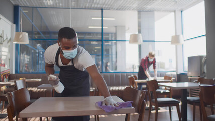 African male worker cleaning table with disinfectant in restaurant during coronavirus outbreak