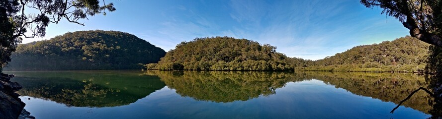 Fototapeta na wymiar Early morning panoramic view of a calm creek with beautiful reflections of blue sky, mountains and trees on water, Cowan Creek, Bobbin Head, Ku-ring-gai Chase National Park, New South Wales Australia