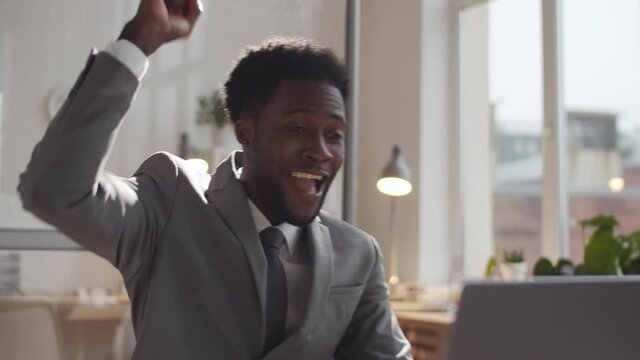 Young afro-american businessman surfing the Internet on laptop at office desk, reading good news, getting excited, smiling and raising arm up while celebrating success
