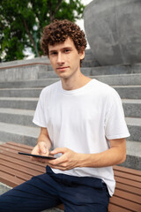 Obraz premium Portrait young European curly man wearing white t-shirt with tablet computer sitting in the park. Freelance outdoor work concept.
