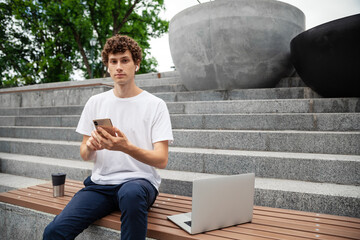 European young man in wireless earphones wearing white t-shirt sitting in a city park and with smartphone and laptop. Freelance work concept outdoors. Empty copy space.
