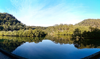 Early morning panoramic view of a calm creek with beautiful reflections of blue sky, mountains and trees on water, Cockle Creek, Bobbin Head, Ku-ring-gai Chase National Park, New South Wales Australia