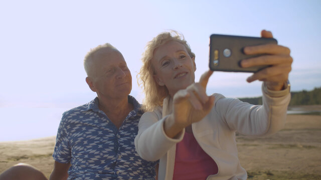 Senior couple taking selfie with mobile phone relaxing on sand at beach