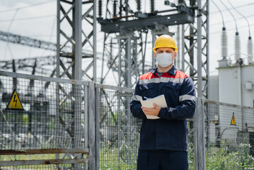 During the pandemic, a masked electrical substation engineer inspects high-voltage equipment....