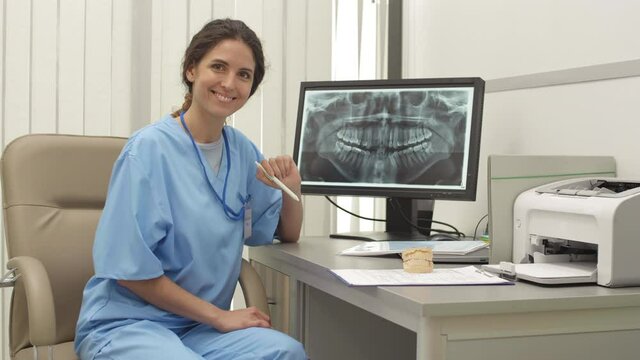 Medium shot portrait of pretty female orthodontist wearing medical overall sitting at desk, examining x-ray image of jaw on computer screen, looking at camera and smiling