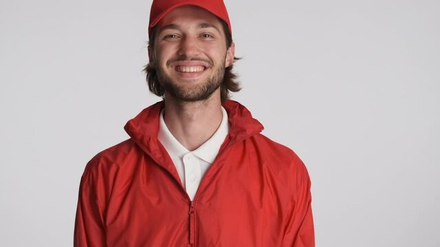 Portrait Of Smiling Delivery Guy In Red Cap And Jacket Happily Looking In Camera And Waving Yes Over White Background