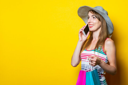 Smiling young woman in hat speaks on the phone, holds packages with purchases on yellow background.