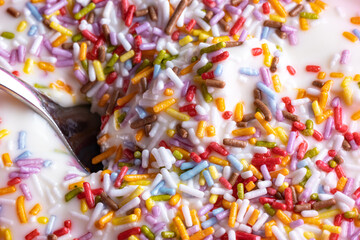 Fototapeta na wymiar Close-up of a mug with yoghurt and colored sprinkles made of sugar, a spoon in the white cream, perfect child dessert for after lunch