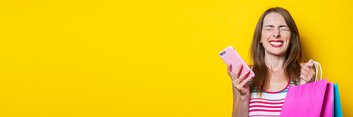 Joyful squinting young girl with phone holding shopping bags on yellow background. Banner.