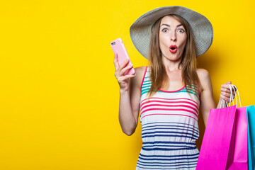 Surprised shocked young girl in hat holding phone and shopping bags on yellow background.