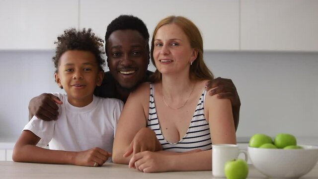 portrait of happy interracial family of three. Spbi couple and mixed race son sit at kitchen and look at camera. teenager son with parents. black dad hugging caucasian mom