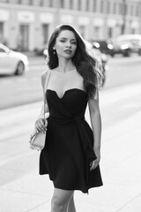 Elegant woman in black dress wearing sunglasses and beige handbag walking city street at sunset....