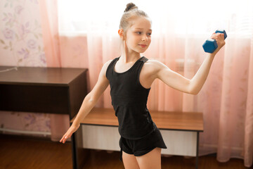 Fototapeta na wymiar beautiful young teenage girl in sportswear is engaged in gymnastics at home with dumbbells
