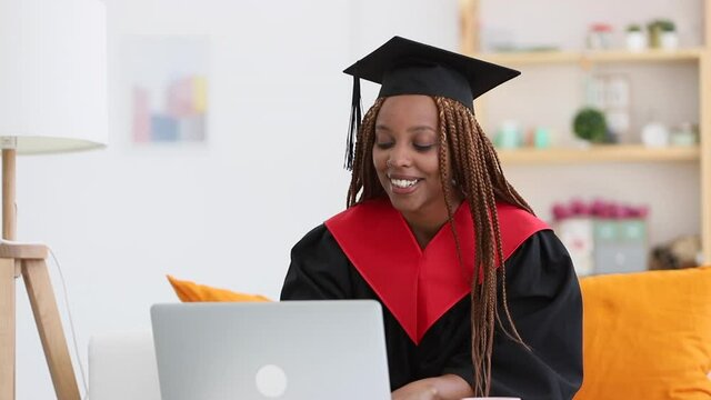 Young female student graduate talking in online chat at table with laptop at home spbi. African American woman has conversation with smile and claps hands, sits in front of computer screen at interior