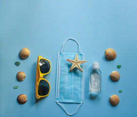 Summer 2020 concept. Travel and protection against coronavirus and other infections. Protective disposable mask, sunglasses, antiseptic, starfish and seashells on blue sunlit background. Copy space
