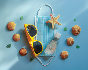 Summer 2020 concept. Travel, vacation and protection against coronavirus and other infections. Protective disposable mask, sunglasses, antiseptic, starfish and seashells on blue sunlit background