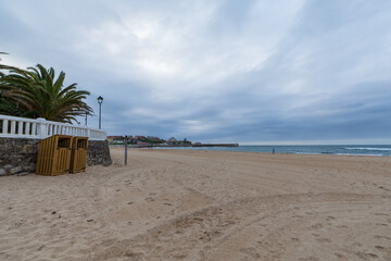 View of beach without people, a cloudy sunset, with palm trees, wall with white fence and wooden huts, horizontal, in Comillas, Cantabria, Spain