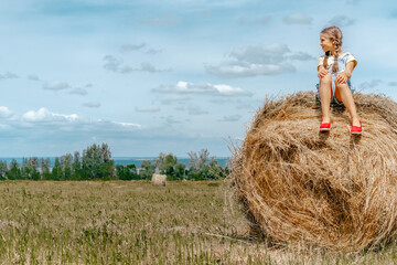 Naklejka na ściany i meble Little girl child in denim overalls sitting on bales of straw, walking in nature in the village