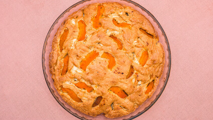 A large, round baked apple and apricot pie stands in a glass form on a table with a tablecloth.