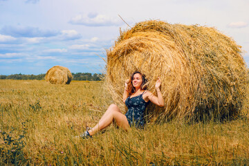 Naklejka na ściany i meble A stylish girl is resting on a bale of hay in a summer field. Portrait of a young sensual woman resting on a haystack, calm moment. Rural slow life