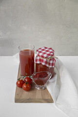 Tomato products are on a white table. This is tomato juice in a glass, sauce, ketchup, tomatoes in their own juice in a jar, next to there are fresh ripe tomatoes on a branch
