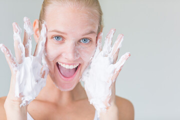 Facial skin care. Pretty playful female model covered in soap foam washing her face with cleanser cream and smiling at the camera. Happy woman isolated against a white background.