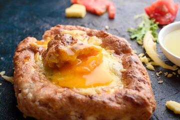Two fresh and delicious khachapuri of weed grain flour and cottage cheese, egg, sulugini cheese, vegetables, oil, cutlery on a dark background.