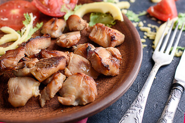 Pieces of baked chicken fillet in spices, fresh vegetables, oil, lettuce, cutlery, round plate on a dark table.