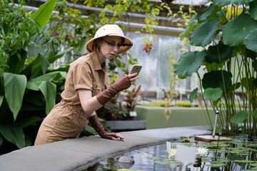 Woman botanist dressed in safari style in greenhouse. Naturalist in khaki clothes exploring aquatic tropical plants in glasshouse, takes a photo on smartphone