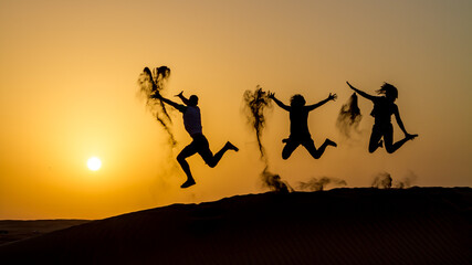 Silhouette of happy traveling people jumping on sand dune and throwing sand in the air in golden sunset hour.