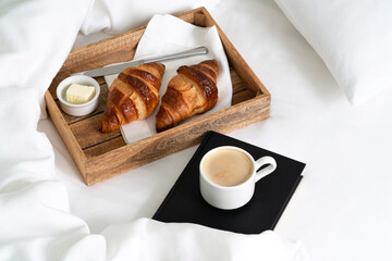 Breakfast in bed with two croissants and butter on the wooden tray, a cup of coffee and book on the white sheet with pillow and blanket. Instagram style.