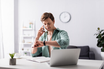 Selective focus of man checking time while talking on smartphone near laptop and notebook on table, concept of time management